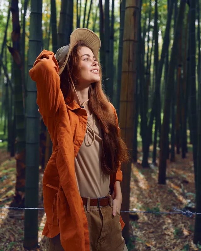 Woman in bamboo forest wearing a straw hat and orange coat, smile and gaze upward in warm sunlight. Portrait of a relaxed traveler with long hair, casual outfit and natural green scenery.