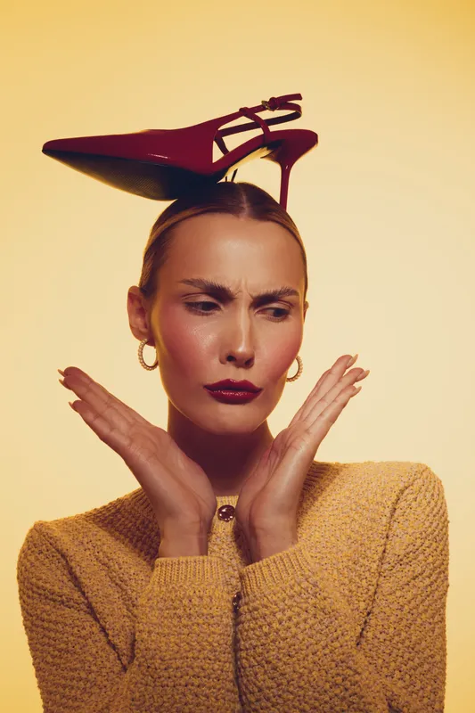 Fashion model with red high heel shoe balanced on head, wearing yellow sweater and gold earrings. Studio portrait with warm lighting and expressive facial expression. Concept of style and creativity.