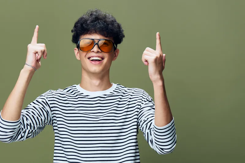 smiling man wearing sunglasses and striped shirt joyfully pointing upwards against green background with playful expression and casual style