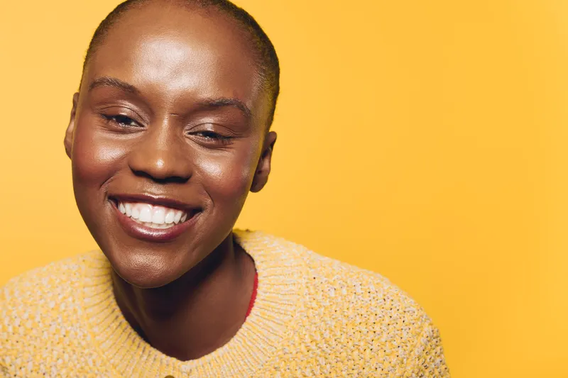 Smiling African American woman with dark skin wearing a yellow sweater poses against a bright yellow background. Concept of happiness, beauty, confidence, and positive emotions.