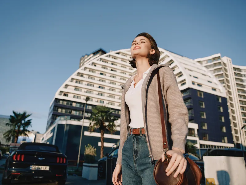 A confident young woman stands outdoors in a sunny urban setting, smiling with optimism as she carries a leather bag, conveying independence and modern, dynamic style.