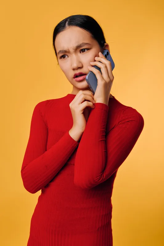 Young woman in red sweater holding phone to ear with worried expression standing against yellow background looking aside. Communication, concern, technology and emotion in portrait.