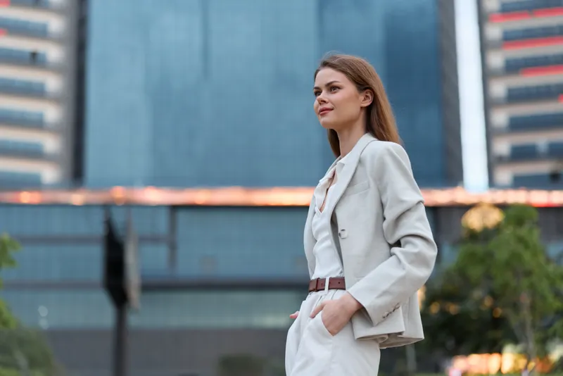 Confident business woman in elegant suit standing outdoors in urban modern setting with buildings, looking thoughtful and professional