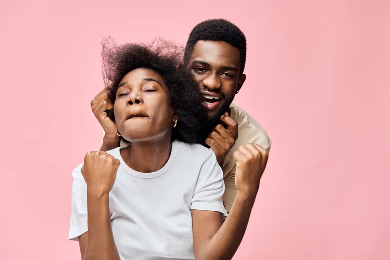 Joyful couple celebrating together with enthusiastic expressions against a pink background, showcasing diverse features and relaxed casual style, emphasizing happiness and connection