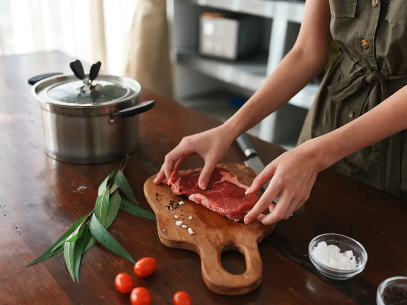 Женщина на кухне готовит ужин из мяса, овощей и специй на деревянном столе на фоне стильной кухни, крупный план рук