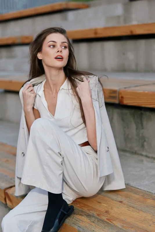 stylish woman in light autumn outfit sitting on wooden stairs outdoor with contemplative expression and modern casual urban style