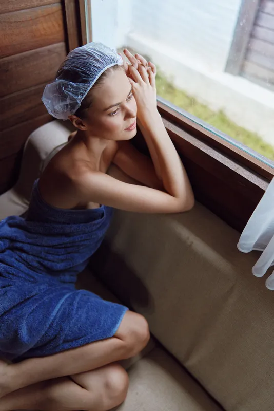Relaxed woman with towel and shower cap sitting by the window, enjoying peaceful moment indoors with soft natural light and cozy wooden interior atmosphere.