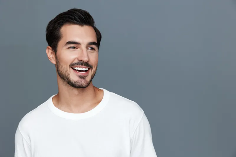 Smiling young man wearing a casual white t-shirt looks camera with a charming smile on a simple solid grey background, showcasing a friendly lifestyle concept.