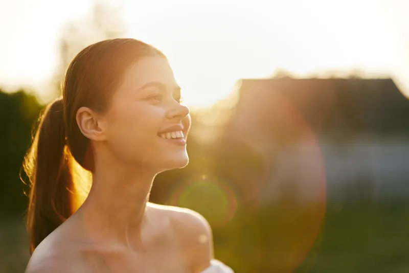 Young woman smiling with sun flare, enjoying nature during sunset, wearing a strapless top, exuding happiness and serenity in a warm, golden landscape