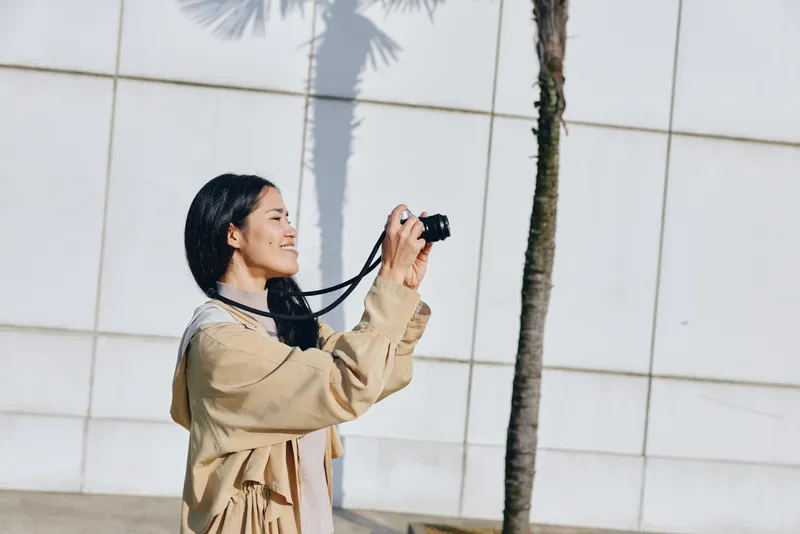 Young photographer capturing scenery with a camera during daytime, wearing a beige coat, smiling as she shoots outdoors beside a palm tree and a sleek urban wall