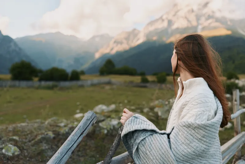 Woman gazing at snowcapped mountains from a wooden railing, wrapped in a cozy sweater. Scenic outdoor portrait conveys calm, nature, and solo hiking vibes in wide open scenery.