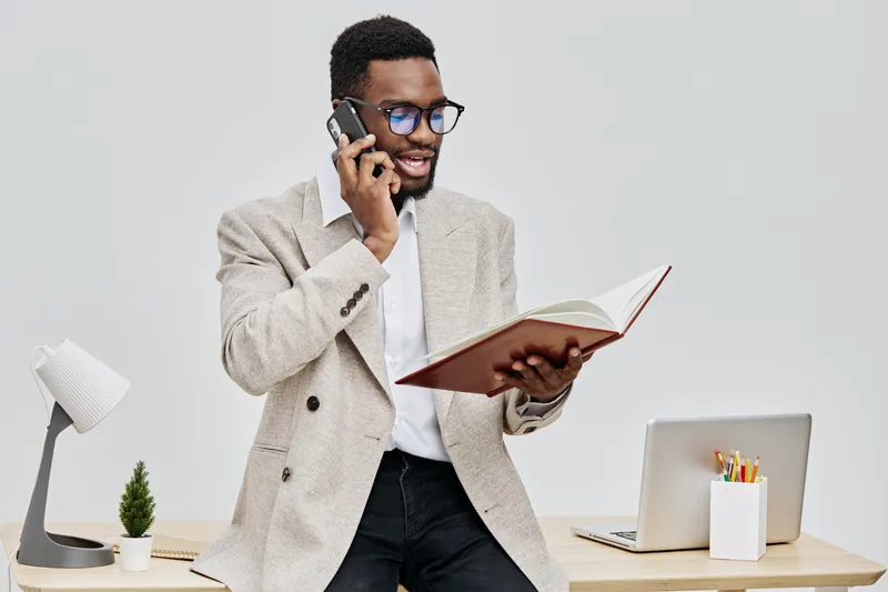 Smiling man talking on mobile phone while holding an open notebook in his hand, standing in modern office with a laptop and desk accessories, dressed in smart casual clothing. People communication con