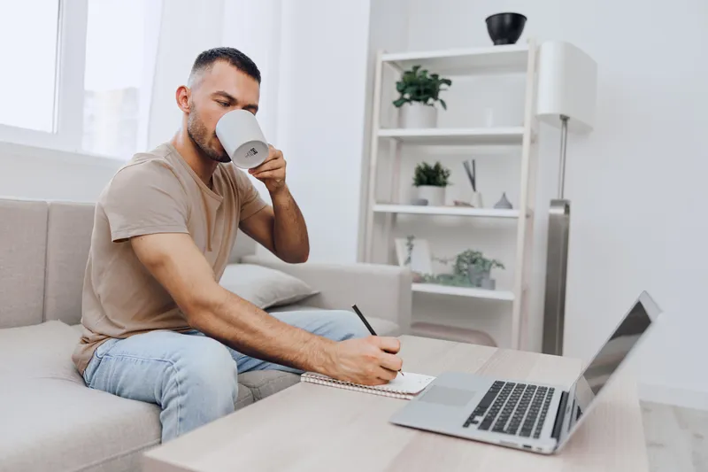 A focused man enjoying a cup of coffee while working on his laptop at home. He takes notes in a notepad, emphasizing productivity and comfort in a modern setting.