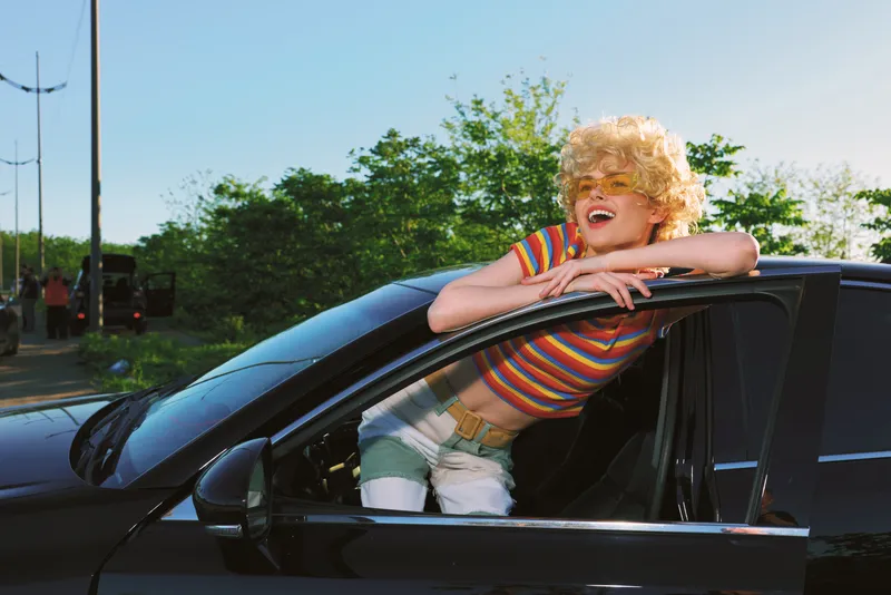 Joyful woman leans out of a sleek black car on a sunny day, wearing a striped shirt and blond wig, capturing a playful retro lifestyle moment with bright outdoor colors and carefree vibe. The scene