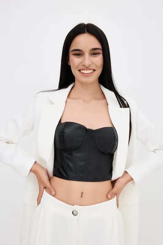 smiling woman wearing black corset top and white blazer with white pants against plain white background, cheerful and modern fashion style.