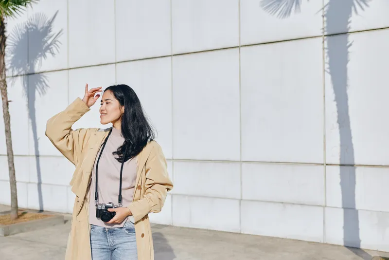 Woman in beige coat adjusts her pose while holding a camera, standing outdoors beside a white wall under bright sunlight, creating vivid shadows and a candid fashion moment