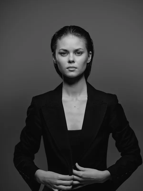 Black and white portrait of a serious woman wearing a blazer with minimal makeup, posing against a dark background.