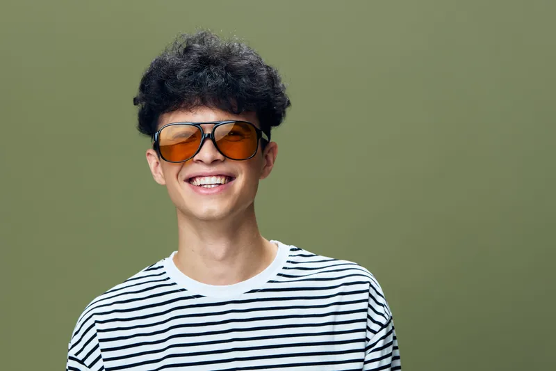 smiling young man wearing striped t-shirt and sunglasses with curly hair on green background, casual style and happy expression