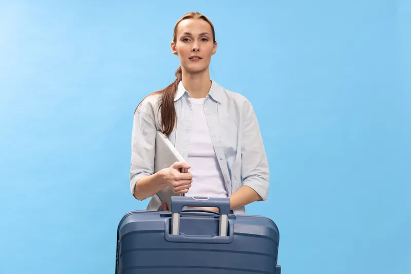 Young woman holding laptop and blue suitcase stands on blue background. Business travel concept with casual outfit and confident expression, isolated studio portrait of traveler or commuter.