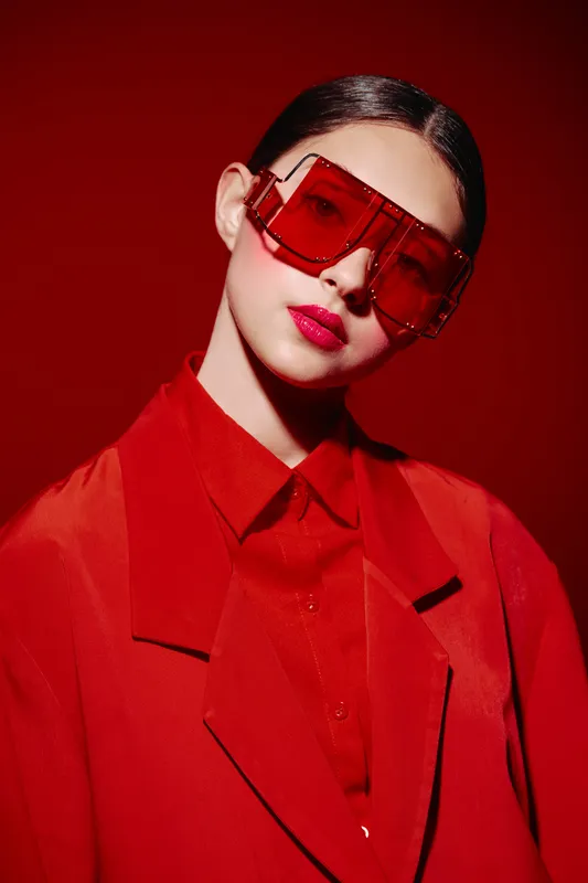 Fashionable woman wearing oversized red sunglasses and a red suit poses against a vibrant red background. The creative beauty shot highlights bold color and modern stylish expression.