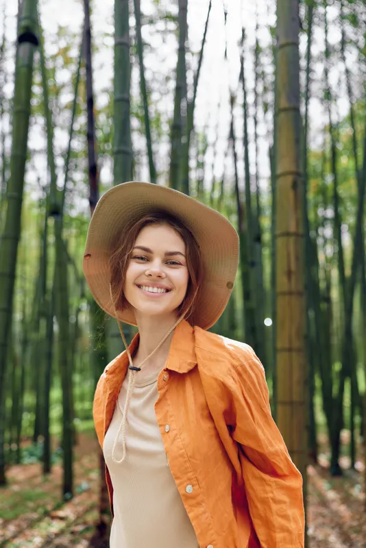 Woman smile hat bamboo forest portrait nature happy in orange jacket and straw sunhat, candid outdoor portrait of a joyful female traveler enjoying green trees and serene natural vibe