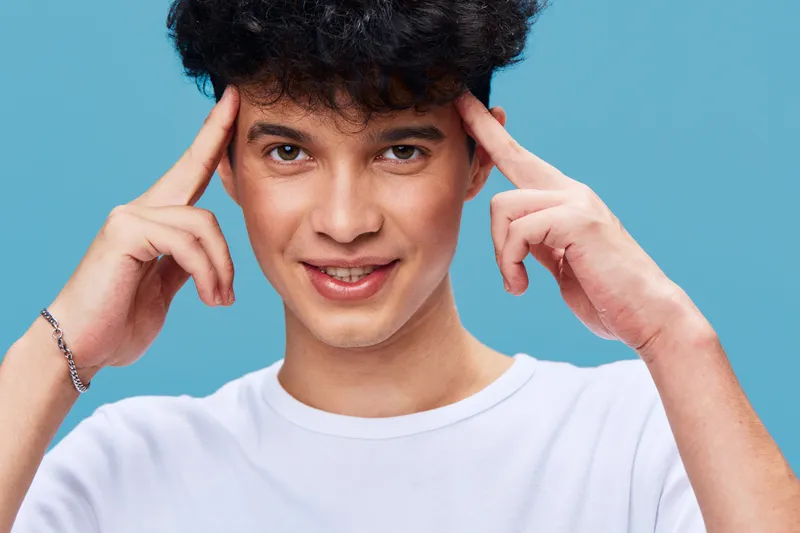 smiling man with curly hair wearing white t-shirt, gesturing with fingers on temples against blue background, casual and youthful expression