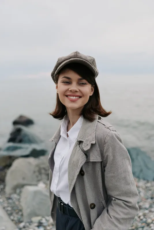 Smiling young woman in vintage outfit stands on rocky beach with calm sea background. She wears a patterned coat and matching cap, expressing happiness and casual style outdoors.