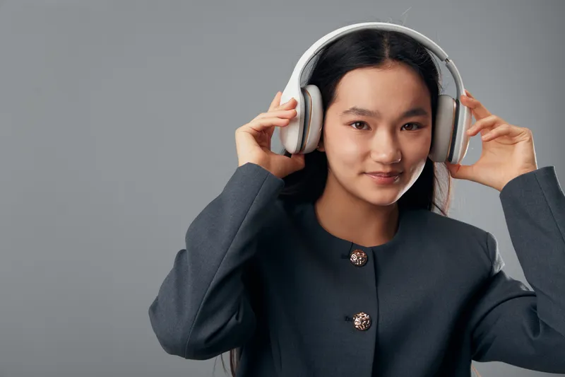 Young woman with headphones smiling and listening to music or audio content on gray background. Portrait of female enjoying sound with hands adjusting headset in casual style.