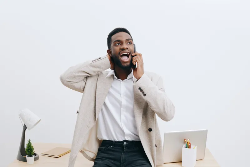 Smiling man talking on phone while standing at modern office desk, dressed in smart casual outfit, happy expression, energetic mood, professional workspace, indoors, corporate environment.