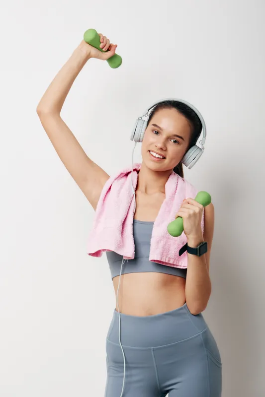 Female fitness enthusiast lifting dumbbells with headphones and a towel, showcasing athletic wear in a bright studio. Dynamic workout moment highlighting strength, focus, and healthy lifestyle in a