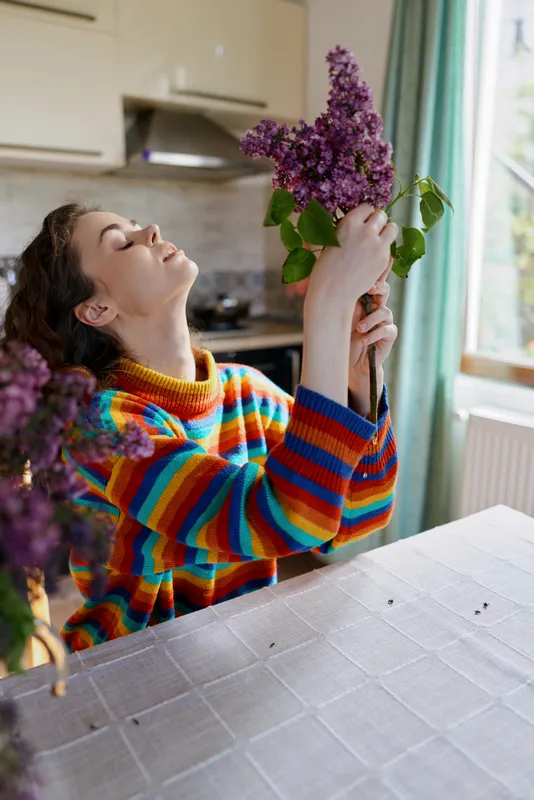 A woman in a vibrant striped sweater adjusts a lilac bouquet on a kitchen table, bright natural light, home setting. Focus on everyday joy, self care, and simple domestic moments.