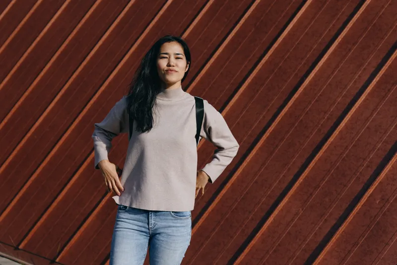 Woman with backpack posing against a warm wooden wall, casual fashion and confident stance, outdoor street style, daylight lighting, urban backdrop, modern wardrobe inspiration.