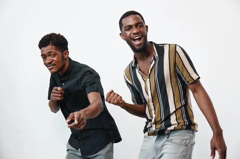Two young black men with dark skin posing and smiling on white background. Casual clothing, joyful expressions, friendship and happiness captured in studio portrait.