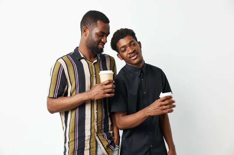 Two black men enjoying coffee together, sharing laughter in a light and cheerful atmosphere. A vibrant moment captured in a simple, modern setting.