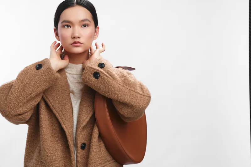 asian woman adjusting a cozy coat in a clean studio setting, she gazes forward with calm confidence, suggesting a ready style for everyday city life and professional presence.