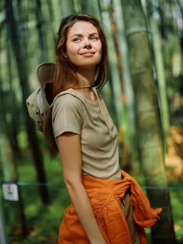 Woman bamboo forest smiling portrait, wearing sun hat and beige top with orange jacket tied at waist, outdoor travel scene in nature with peaceful sunlight and relaxed mood.