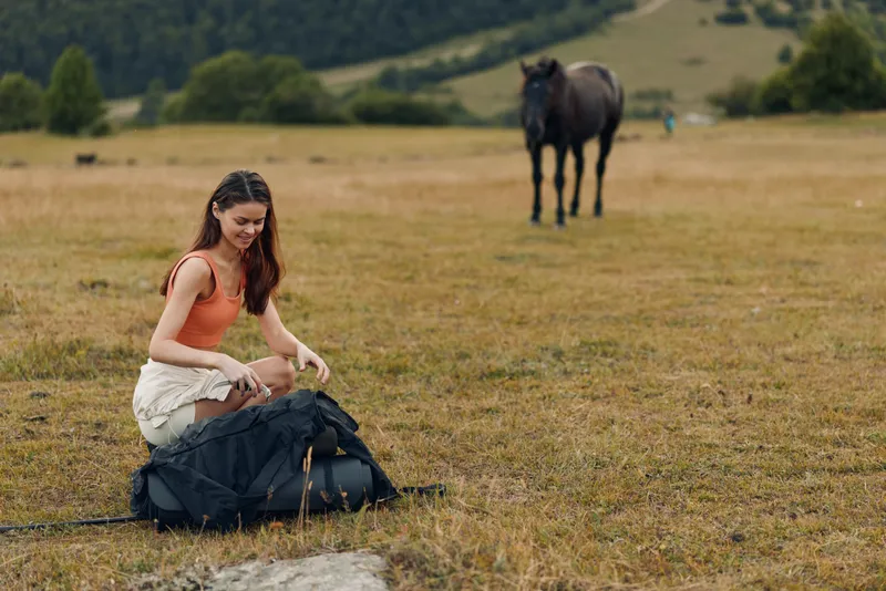 A young woman kneels on a grassy field, packing a backpack. In the background a calm horse stands near rolling hills, creating a serene, candid outdoor moment.