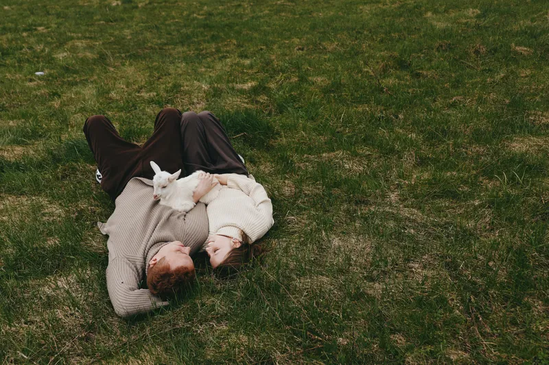 Romantic couple lies on grass, embrace and whispers, outdoor affection on a tranquil lawn, warm sweaters and a cozy mood, candid moment of closeness in natural light