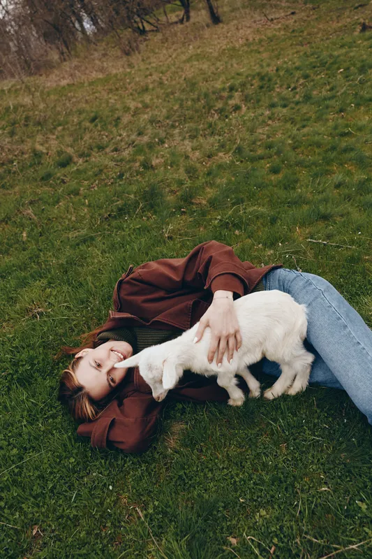 Young person lying on grass with playful goat kid, enjoying a relaxed outdoor moment, casual clothing, gentle interaction and affection in a tranquil park