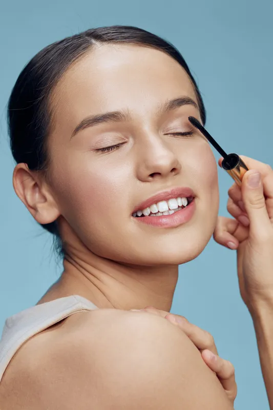 Young woman applying mascara with a joyful expression against a light blue background, showcasing beauty and makeup concepts in a bright, fresh style