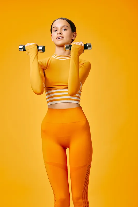 Woman in bright orange athletic wear lifts dumbbells in a bold studio, showcasing fitness training and strength. Vibrant orange background mirrors outfit for a cohesive sport portrait focused on