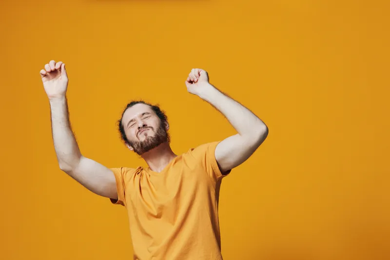 Joyful man celebrating with raised arms against a vibrant orange background, wearing a casual orange t shirt, expressing happiness and positivity in a bright, energetic atmosphere