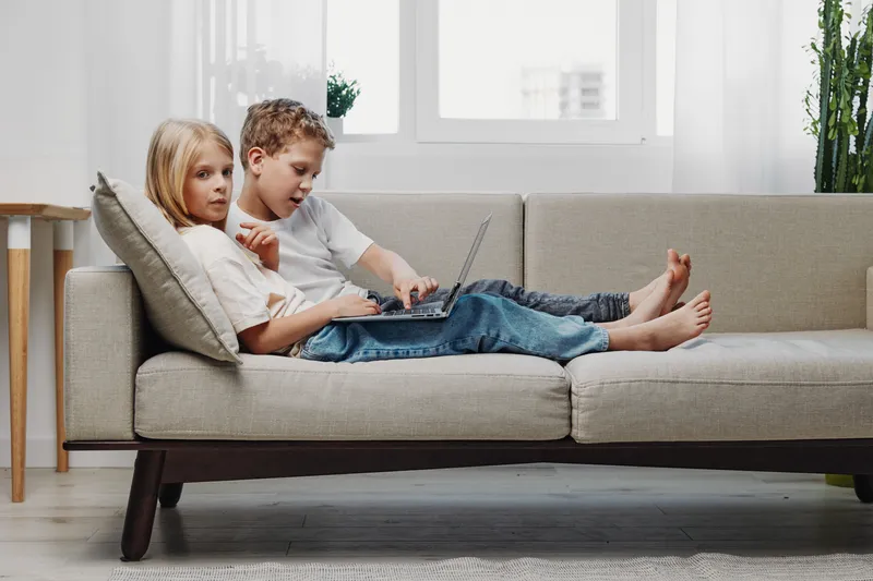 Two children relax on a modern sofa using a laptop together in a bright living room. Casual home scene with focus on technology, leisure, and sibling interaction in natural daylight.