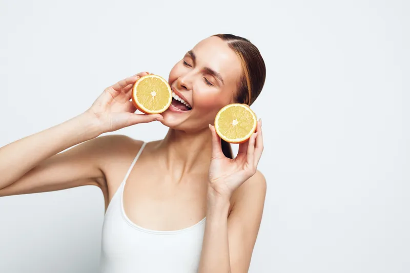 Cheerful woman holding fresh orange halves near mouth, bright citrus tones, white tank top, clean studio backdrop, playful healthy moment, natural glow, vibrant texture and clear color