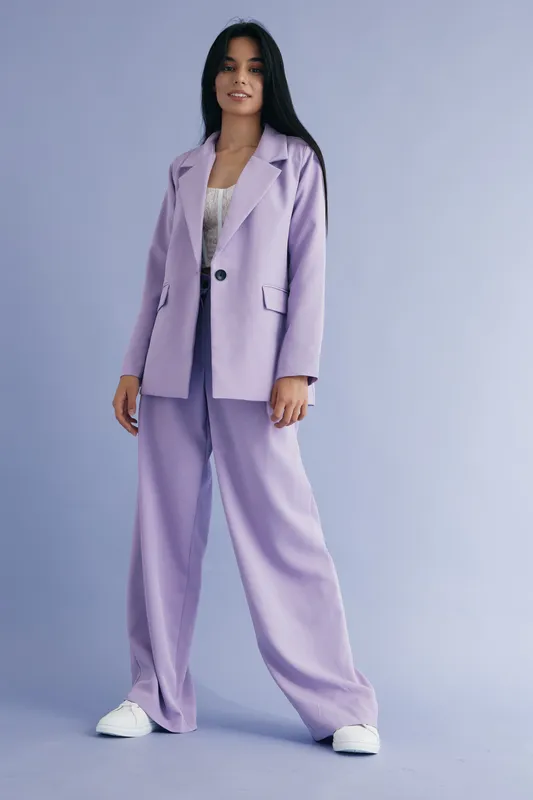 Young woman wearing a lavender pantsuit stands confidently against a soft purple background, showcasing modern fashion with a relaxed yet stylish pose and casual white sneakers.