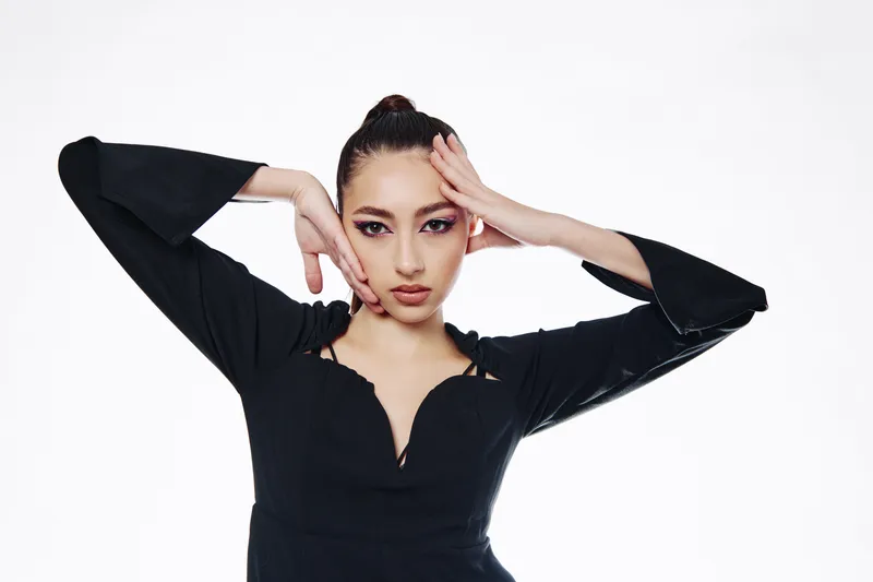 Fashion model with sleek bun wears a black stylish top posing with hand on face against white background. Creative beauty concept with confident expression and sharp makeup.