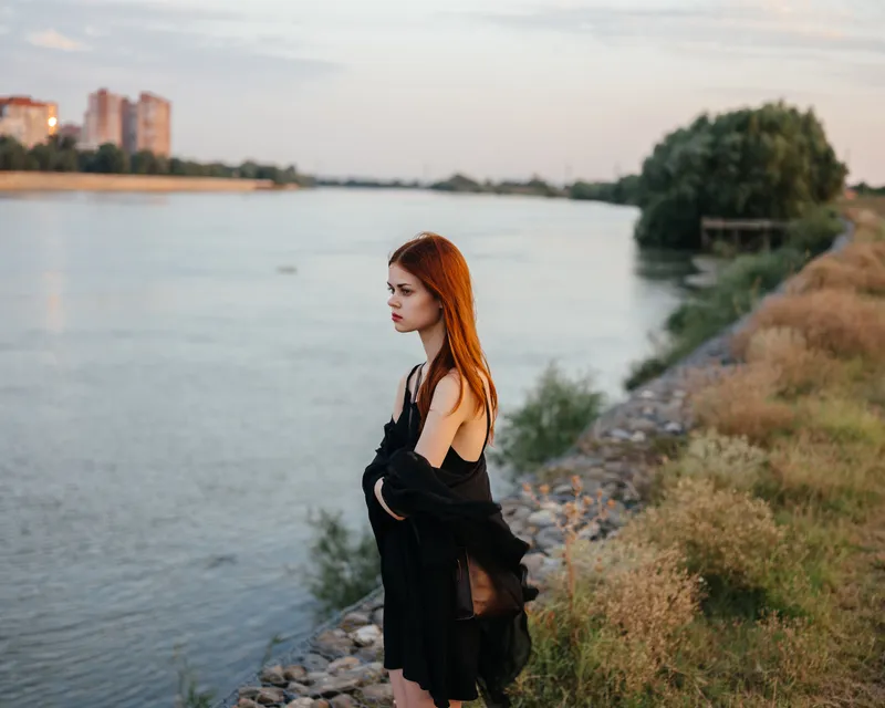 Рыжеволосая женщина в черном платье стоит у реки на природе