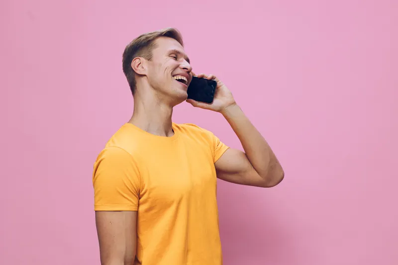 Smiling man communicating on mobile phone with happy expression, wearing casual yellow t-shirt, standing against pink background. Bright, cheerful, relaxed mood. People lifestyle concept.