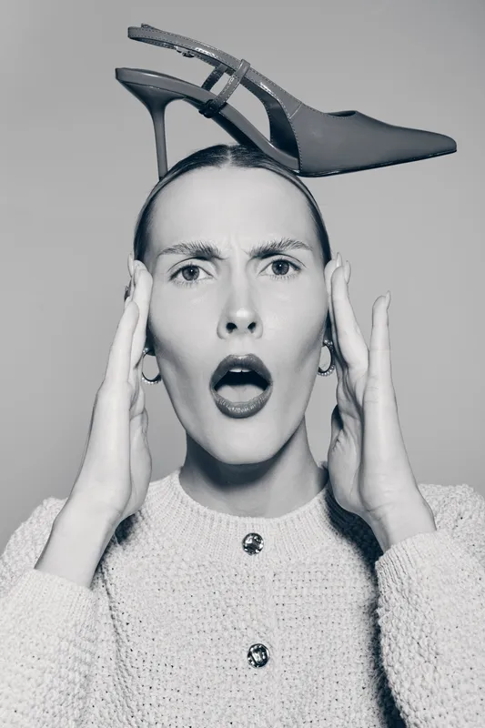 Surprised white woman with pale skin holds black high heel shoe on head in studio. Concept for fashion, creativity and unusual style.