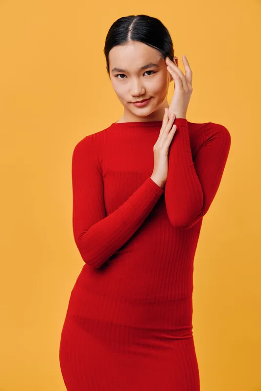 Young woman in red dress posing against orange background with hands near face smiling gently. Female model with dark hair and clear skin wearing fitted long sleeve outfit in studio.
